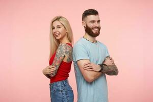 Happy attractive tattooed young people keeping thier hands folded and looking cheerfully ahead with pleasant smiles, standing back to back over pink background photo