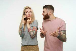 Studio photo of young long haired blonde lady looking pensively upwards while having phone talk, standing over white background with bewildered bearded tattooed male