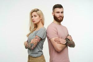 Studio photo of pretty pair of young people crossing hands on chests and looking seriously at camera with folded lips, standing over white background