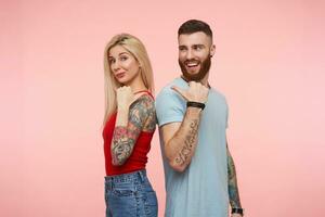 Horizontal shot of positive lovely couple standing back to back and keeping hands raised while pointing on each other with thumbs, isolated over pink background photo