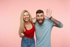 Horizontal shot of lovely joyful pair of young people fooling and laughing while spending nice time togehter, isolated over pink background in casual wear photo