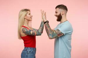 Studio photo of lovely young positive couple dressed in casual clothes smiling to each other and keeping palms raised while giving five, posing over pink background