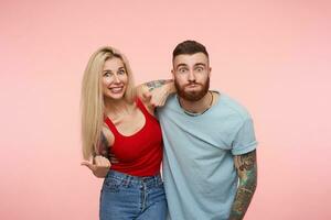 Positive young pretty couple with tattooes embracing each other while making funny face and looking excitedly at camera, standing over pink background photo