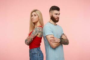 Serious tattoed pair of yuong people crossing hands on chests and frowning sadly eyebrows, wearing casual clothes while standing over pink background photo