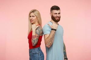 Displeased young pretty tattooed people showing on each other with raised hands and frowning discontentedly faces while looking at camera, isolated over pink background photo