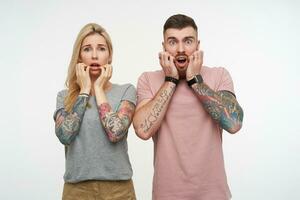 Shocked young pretty tattooed couple holding raised hands on their cheeks while looking amazedly at camera with wide eyes opened, isolated over white background photo