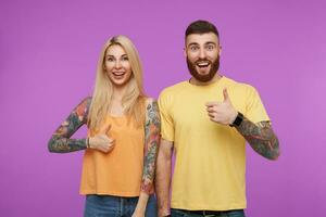 Cheerful lovely tattooed pair of young people showing raised thumb while looking happily at camera with wide smiles, standing against purple background photo