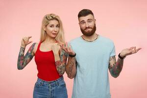 Indoor photo of positive young pretty bearded man shrugging with raised hands while standing with his confused blonde girlfriend showing small size over pink background