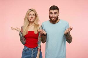 Horizontal photo of young pretty couple with tattooes biting worringly underlips and wrinkling foreheads while posing over pink background with raised hands