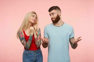 Cute young long haired blonde female with tattooes folding raised hands while asking something from her pretty bearded tattooed friend while being isolated over pink background photo
