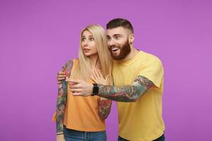 Surprised young lovely people in orange casual clothes rounding wonderingly their blue eyes while looking aside, standing against purple background photo