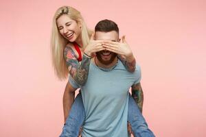 Studio photo of young pleasant looking joyful blonde female sitting on back of her handsome bearded boyfriend and covering him eyes with raised palms, isolated over pink background