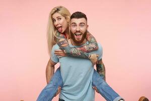 Funny shot of young lovely tattooed couple dressed in casual clothes expressing their positive emotions and making faces while standing over pink background photo