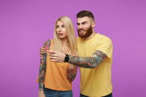 Confused young tattooed bearded male grimacing his face while pointing aside with forefinger and hugging his frightened pretty long haired blonde girlfriend, isolated over purple background photo