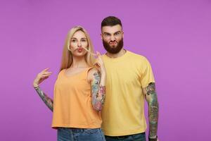 Indoor shot of young attractive tattooed couple grimacing their faces while fooling, spending nice time together while posing over purple background photo