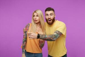 Excited young tattooed handsome man with beard looking surprisedly at camera and embracing his confused long haired blonde girlfriend while posing over purple background photo
