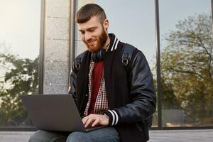 Outdoor shot of handsome red bearded young guy, sitting on the street putting the laptop on lap, smiling and chatting with his friends. photo