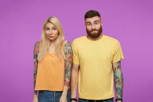 Puzzled pretty pair of young people with tattooes twisting their mouths and shrugging with hands down while being isolated over purple background in casual wear photo