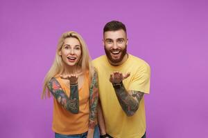 Portrait of young happy couple with tattooes keeping their palms up while looking cheerfully at camera with wide smiles, isolated over purple background photo