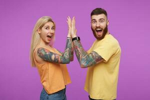 Astonished young attractive tattooed couple clapping raised palms and looking excitedly at camera with wide mouths opened, isolated over purple background photo