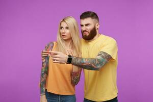 Bewildered young bearded brunette man hugging his afraid blonde tattooed girlfriend while showing ahead and grimacing his face, posing over purple background photo