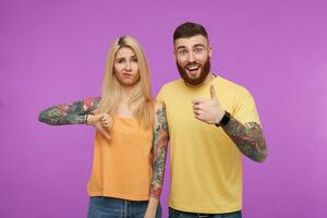 Horizontal shot of young pretty tattooed couple in casual wear keeping their hands raised while gesturing and expressing different emotions, isolated over purple background photo