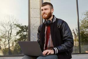 Outdoor shot of handsome red bearded young guy, sitting on the street putting the laptop on lap, looking into the distance and planning a trip to the mountains. photo