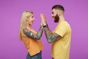 Side view of young cheerful tattooed couple looking happily on each other and keeping hands raised while giving five, standing over purple background photo