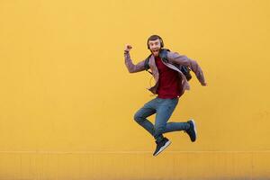 Happy young handsome ginger bearded man jumping like superhero, full of energy, wearing in basic clothes with backpack. Looking at the camera with wide open mouth over a yellow wall with copy space. photo