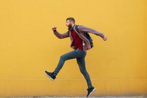 Young attractive ginger bearded man jumping, very fast running, late for work, wearing in basic clothes with backpack. Isolated over yellow wall with copy space. photo