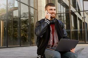 Outdoor shot of cool red bearded young man, sitting on the street putting the laptop on lap, has telephone conversation with frined, enjoys excellent cellular communication and free wi-fi. photo