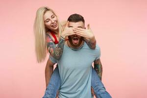 Attractive young long haired positive woman making surprise to her lovely tattooed bearded boyfriend and closing him eyes with raised hands, isolated over pink background photo