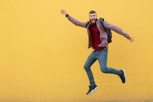 Happy smiling young attractive ginger bearded man jumping, wearing in basic clothes with backpack. Looking at the camera with wide open mouth over a yellow wall with copy space. photo
