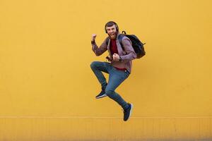 Smiling young attractive ginger bearded man jumping,happy success, wearing in basic clothes with backpack. Looking at the camera over a yellow wall with copy space. photo