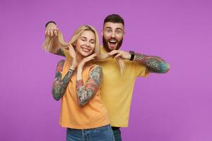 Horizontal photo of young lovely tattooed couple dressed in orange casual clothes being in nice mood while having fun together, standing over purple background