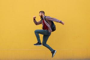 Young attractive ginger bearded man jumping, very fast running, hurry to the store for sale, wearing in basic clothes with backpack. Isolated over yellow wall with copy space. photo