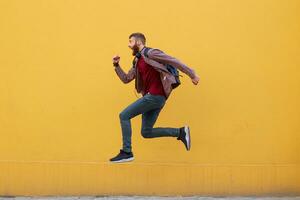 Young attractive ginger bearded man jumping, very fast running, running away from hateful work, wearing in basic clothes with backpack. Isolated over yellow wall with copy space. photo