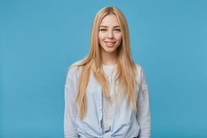 retrato de joven rubia atractivo mujer con largo pelo vistiendo azul camisa y gris camiseta, en pie terminado azul antecedentes con manos abajo, mirando a cámara con encantador sonrisa foto