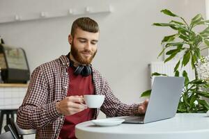 contento atractivo joven jengibre barbado hombre trabajando a un computadora portátil, sentado en un cafetería, vistiendo en básico ropa, sonriente y disfrutar el café. foto