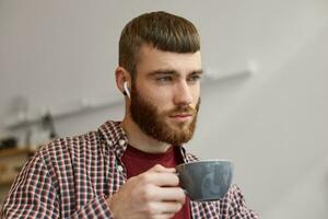 Photo of young attractive ginger bearded man holding a gray coffee cup, think about something and looking away, wearing in basic clothes.