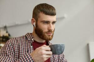 foto de joven atractivo jengibre barbado hombre participación un gris café taza, refleja en planes para mañana y mirando lejos, vistiendo en básico ropa.