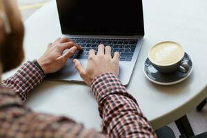 In a cafe on a white table, men's hands are working on the keyboard of the laptop, to his right is a cup of cappuccino. photo