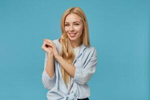 Charming young blonde long haired woman raising folded hands and smiling to camera cheerfully, standing over blue background in casual clothes, demonstrating her white perfect teeth photo