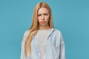 Close-up of offended blonde long haired female wearing blue shirt and grey t-shirt, looking at camera with folded lips and frowning, posing over blue background with hands along body photo