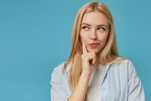 Close-up of beautiful young blonde lady with casual hairstyle looking aside thoughtfully, keeping forefinger on her cheek and smiling cunningly, isolated over blue background photo