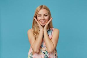 Cheerful pretty blonde long haired lady wearing flowered dress, looking at camera with charming smile and leaning chin on raised palms, isolated over blue background photo