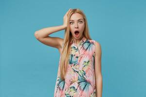 Shocked blonde long haired female wearing romantic dress, keeping palm on her head while posing over blue background, rounding eyes with mouth opened photo