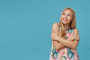 Lovely young female with blonde long hair wearing romantic dress with floral print, keeping hands crossed on her chest and looking aside with charming smile, isolated over blue background photo