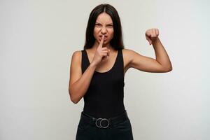 Severe young dark haired female with casual hairstyle frowning viciously her face and showing on camera with forefinger, asking to be quiet while standing over white background photo