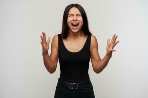 Excited good looking young dark haired woman with casual hairstyle keeping her mouth wide opened while frowning her face with closed eyes, raising emotionally palms over white background photo